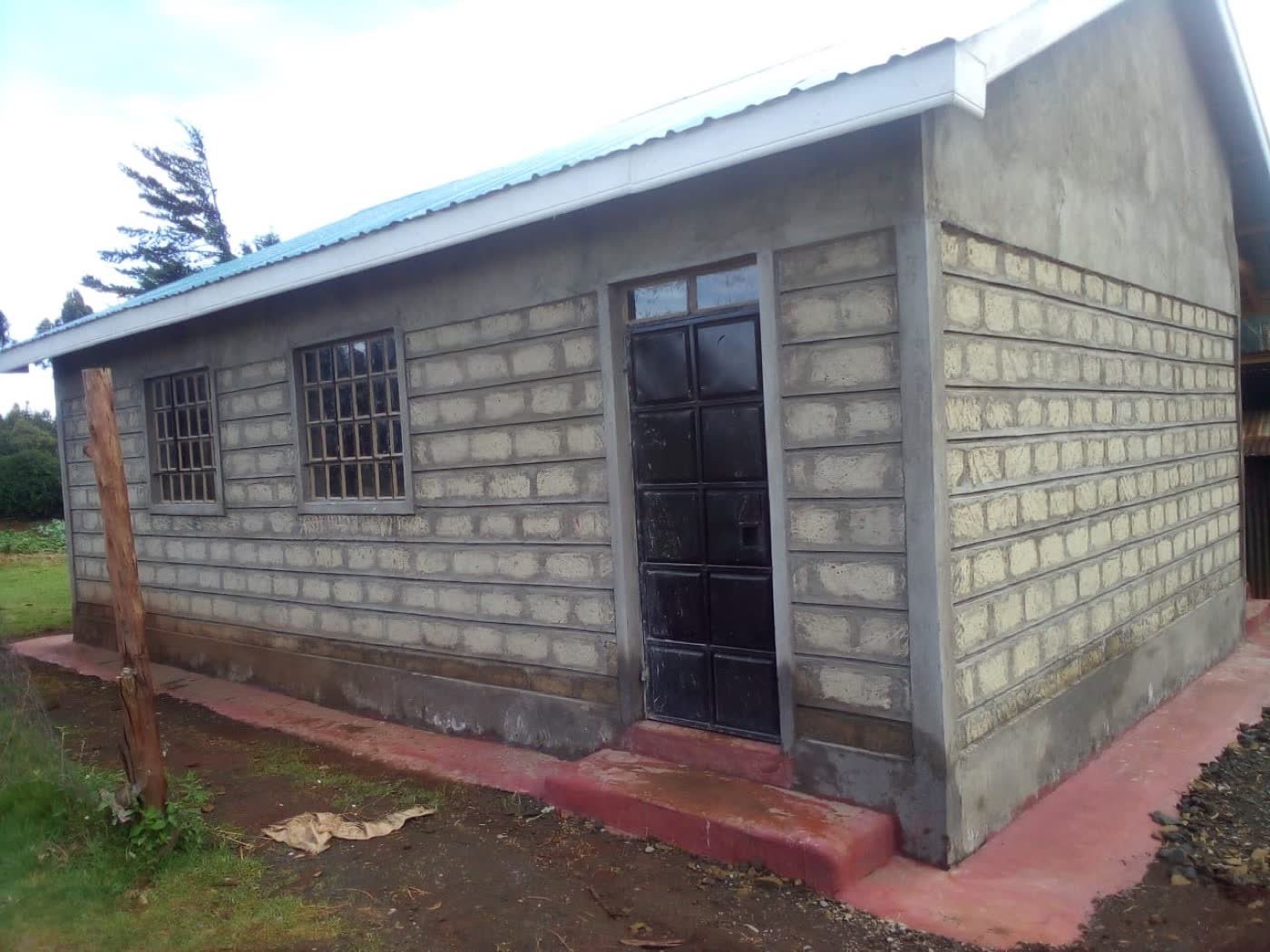 A stone-block school building with a plastered gable, steel door and windows, and a concrete apron