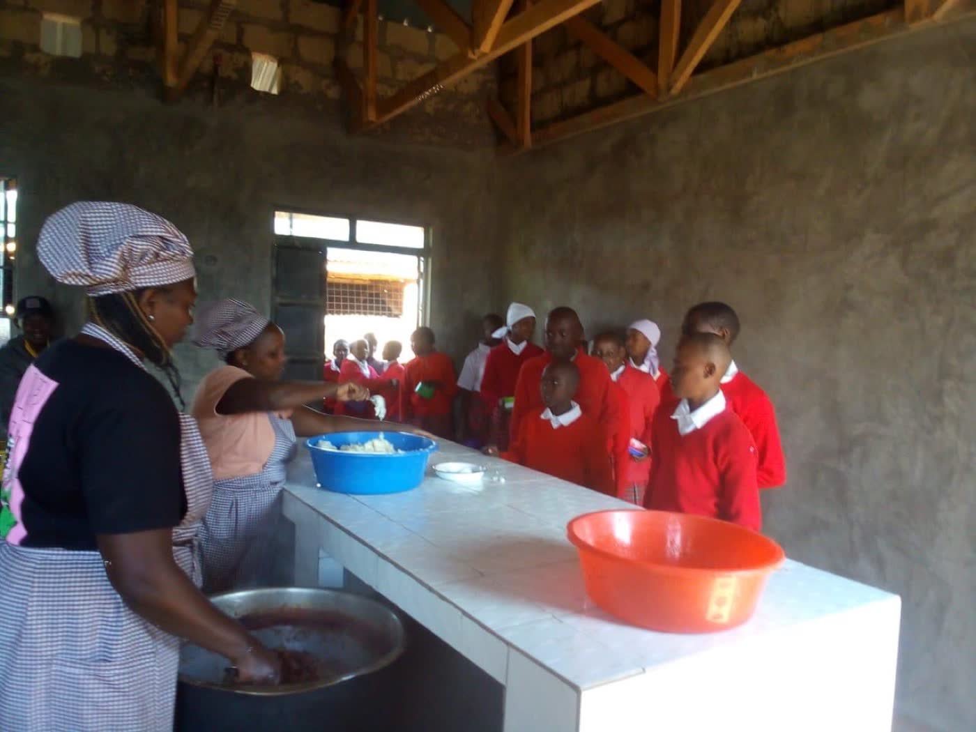 Two cooks in aprons serving food from bowls across the tiled counter to a line of students in red uniforms