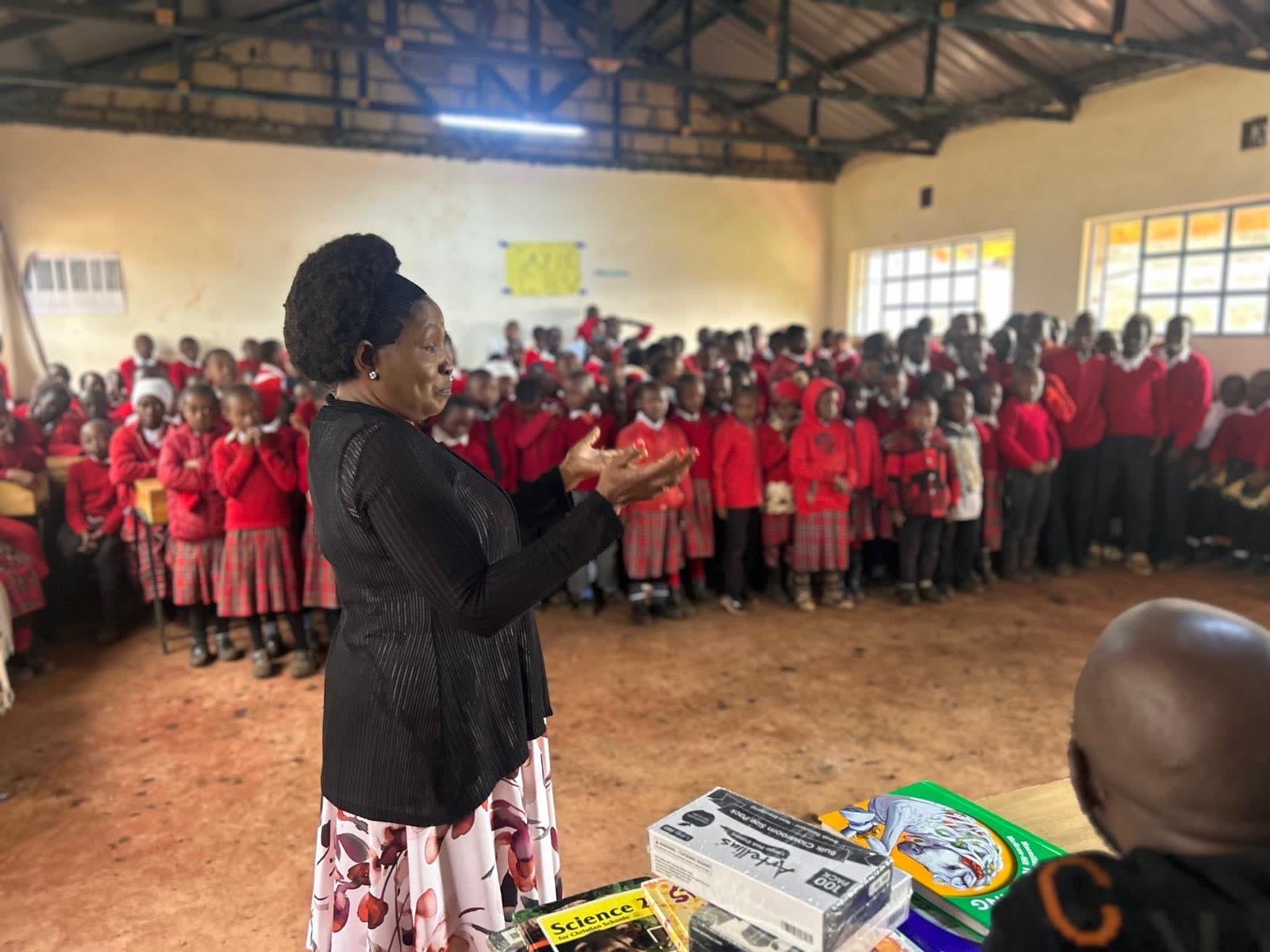 Dr. Naomi Karaya speaking to a hall full of students beside a table of donated books and supplies
