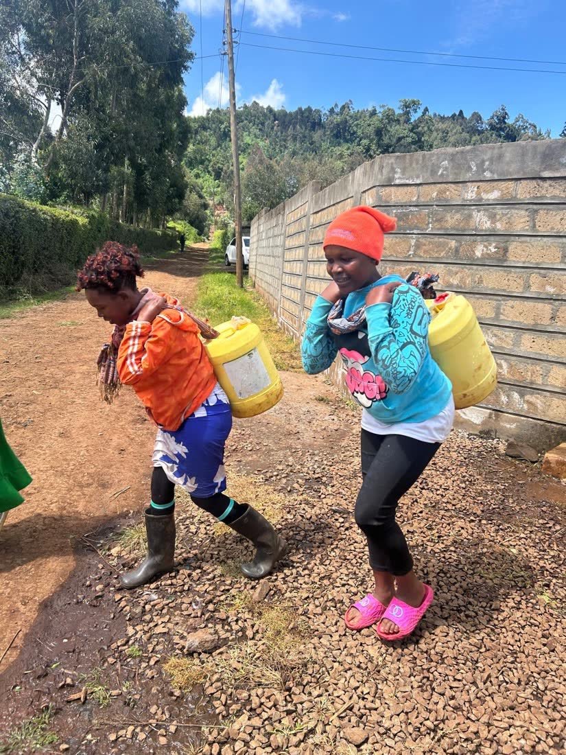 Two young women carrying heavy yellow jerrycans of water on their backs along a dirt road