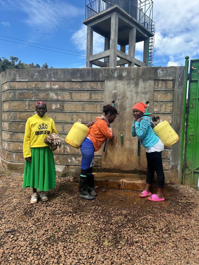 Three women filling yellow jerrycans at a row of taps set into a stone wall beneath an elevated water tank