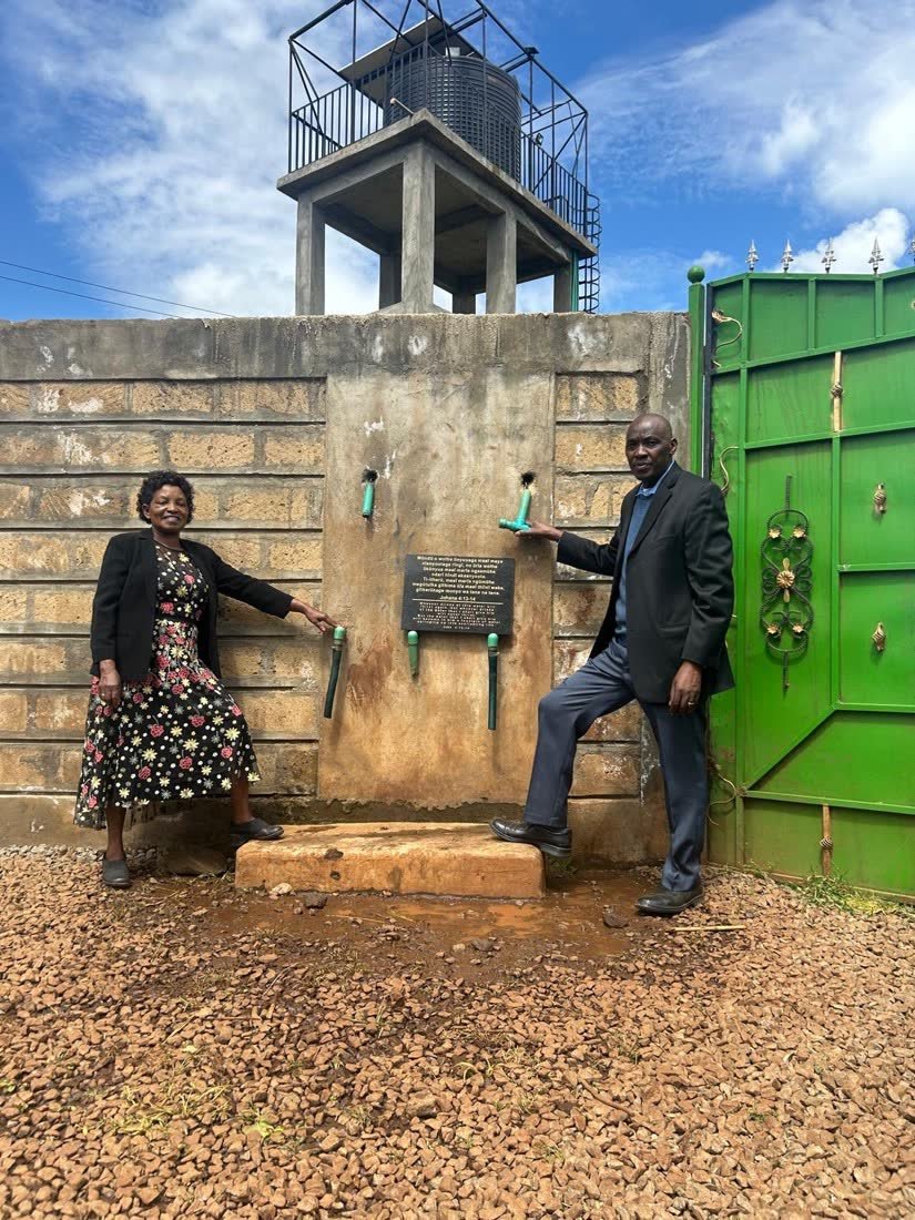 Two people beside the water point wall with its dedication plaque, beneath an elevated tank